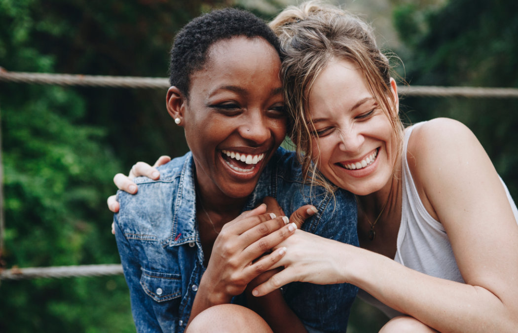 Two women smiling and laughing together outdoors, sitting closely with arms around each other. Green foliage is visible in the background.