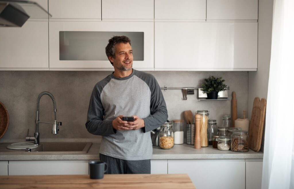 A casually dressed man stands in a bright, contemporary kitchen, looking at his phone with a smile. The counter features jars, cutting boards, and kitchen tools, suggesting everyday home life.