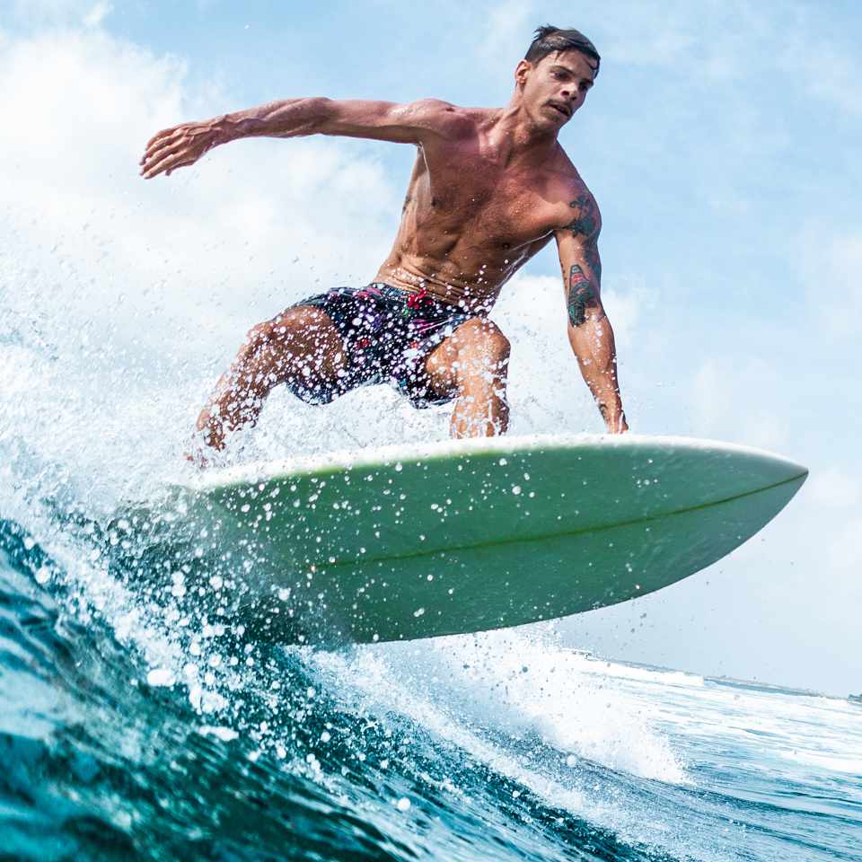 Shirtless man with tattoos surfing a wave on a green surfboard, arms extended for balance with water spraying around him.